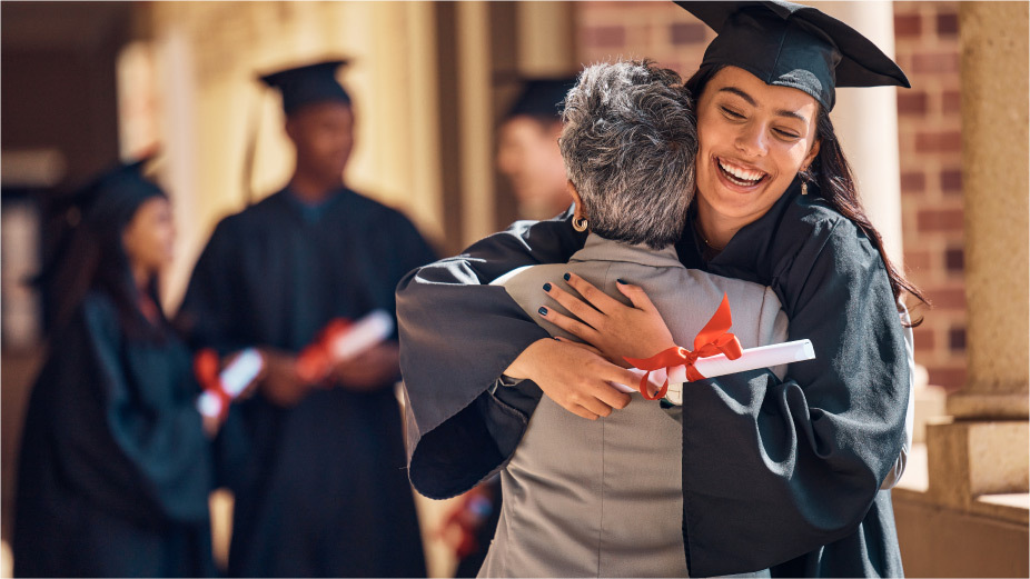 A happy graduate dressed in a cap and gown receives a hug.